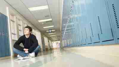 A teen boy sitting in a high school hallway, struggling to manage his workload